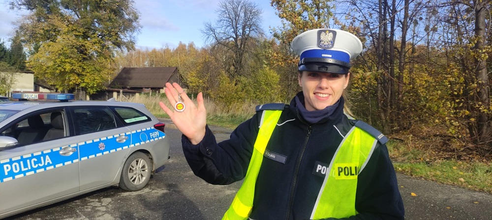Na zdjęciu funkcjonariuszka Policji, która w ręku trzyma brelok rozdawany kierowcom podczas akcji Bezpieczna podróż = Trzeźwe życie. W tle radiowóz.